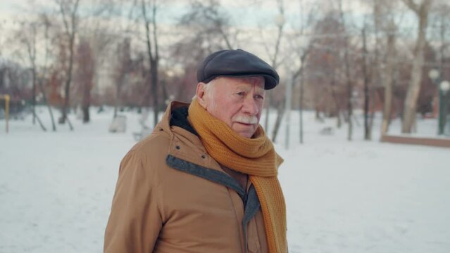 Waist-up of elderly Caucasian man wearing flat cap, jacket and scarf, walking in park in winter