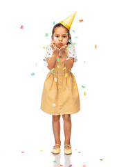 birthday, childhood and people concept - little girl in dress and party hat blowing to confetti over white background