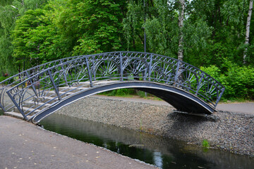 arched metal bridge across water canal in the public park on green trees background