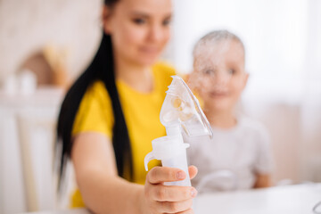 Obraz na płótnie Canvas an inhaler with a nebulizer at home against the background of a little boy and his mother. A child with a viral disease is sitting in his mother's arms. Patient with asthma