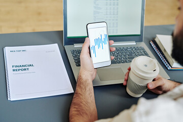 Young male office worker holding glass of coffee and smartphone with financial charts on screen...