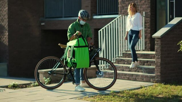 Outdoor man courier in a safety helmet and medical mask delivering food in a paper bag to the customer home by bicycle attractive woman stand outside order coronavirus contact less slow motion
