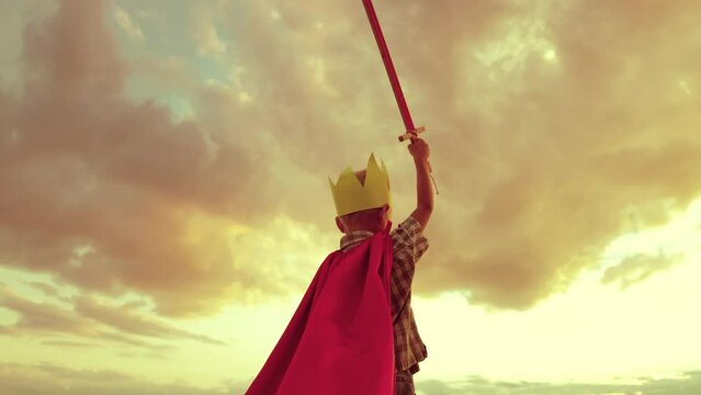 boy plays superhero against backdrop sunset sky. small child kid red cape with sword his hand. child dream kid. boy golden crown plays game. costume children's fantasy happy baby vacation holiday.
