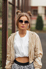 Stylish beauty girl with hairstyle, pink lips and round vintage sunglasses in sporty clothes with windbreaker, t-shirt, and leggings walks on the street .