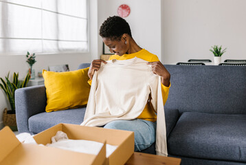 Young african woman receiving and opening a parcel box with a clothing item from an online purchase...