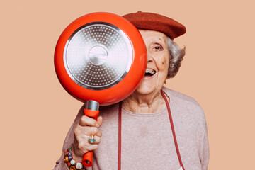 Portrait of a happy smiling elderly grandmother covering half face with a pan at studio over beige...