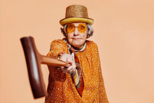 Funny stylish elderly grandmother aiming at camera with a cane at studio over beige background
