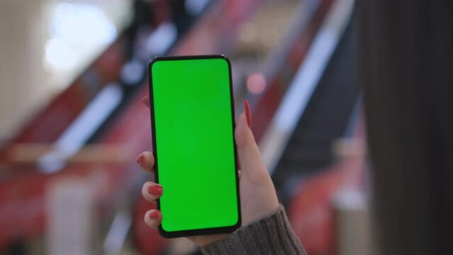 Hands of a young woman holding a smartphone in a shopping mall. A young woman is holding a phone in a shopping mall in front of an escalator with people. Close up