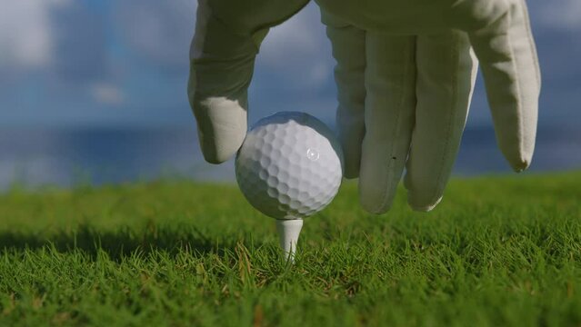 Male hand in a golf glove puts the ball on the field on the background of the blue ocean. Close up