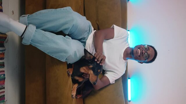 African American guy strokes a dachshund and waves his hand indignantly while watching sport on TV. Slow motion. Vertical video
