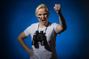 Aggressive blonde woman with a blue-yellow Ukrainian flag on her cheek with binoculars around her neck screams and shows her fist on a blue background.