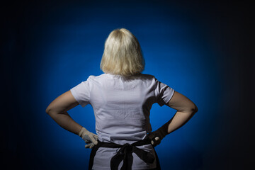 A woman in a black apron and gloves stands with her back to the camera against a dark background.