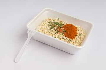 dry instant noodles with a plastic fork, vegetables and hot spices in a container on a white background. fast food concept