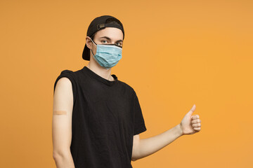 A young man in two medical masks after vaccination with a medical plaster on his hand shows a thumbs up, portrait on a yellow background. The concept of vaccination against coronavirus.