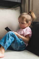 Little girl is sitting on sofa with smart phone. She's watching intently in it.