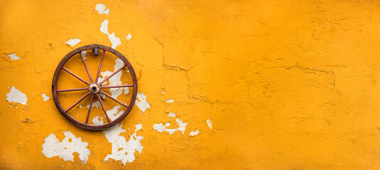 Fototapeta stare koło od wozu powieszone na żółtej obdrapanej z farby starej ścianie obraz