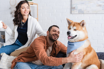smiling man sitting on couch and cuddling akita inu dog near blurred girlfriend with cup.