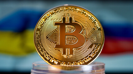 Golden cryptocurrency bitcoin in front of the flag of Ukraine and Russia. Focus is on the coin in the front.