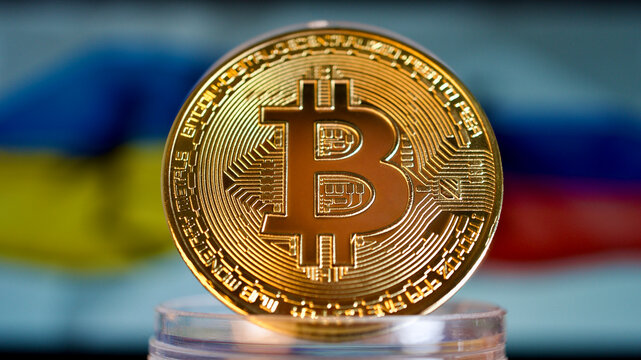 Golden cryptocurrency bitcoin in front of the flag of Ukraine and Russia. Focus is on the coin in the front.