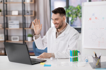 distant education, school and science concept - happy smiling male chemistry teacher with laptop computer having online class and waving hand at home office