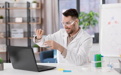 distant education, school and science concept - happy male chemistry teacher in goggles and earphones with laptop having online class and pouring chemical from test tube to flask at home office