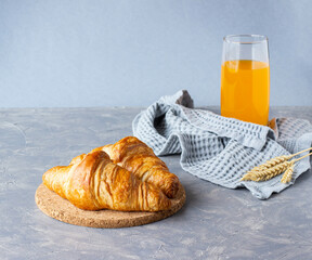 two croissants are lying on a wooden substrate and a glass of orange juice is on the table, on a gray background, the minimalism of a healthy breakfast