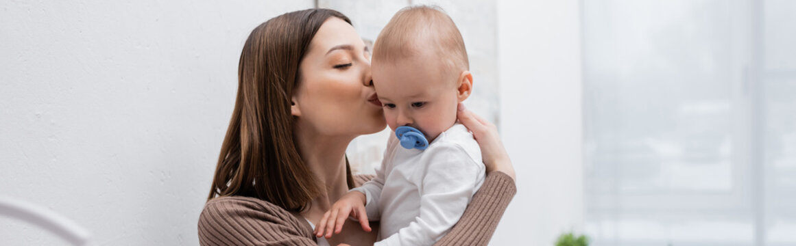 Woman kissing baby son with pacifier at home, banner.