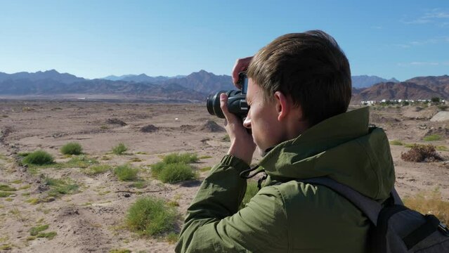 Photographer takes pictures of mountain landscape. Male tourist, traveller photographing arid desert area with rocks. Profile back view. Journalist shoots photos of barren place or video for report