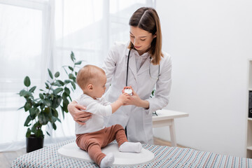 Baby touching stethoscope on doctor while sitting on electronic scale in clinic.