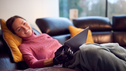 Woman With Prosthetic Arm Using Laptop Relaxing On Sofa At Home