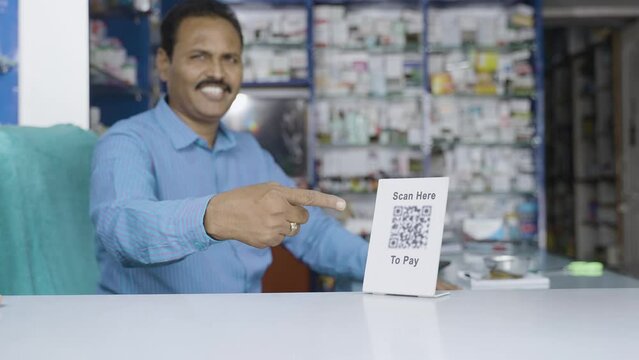 handheld rack foucs shot of Smiling pharmacist showing scan here to pay qr code for payment by looking at camera at store - concept of online payment, small business and technology.