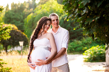 Young happy romantic pregnant white couple in white shirt, dress, kissing, hugging, touching belly in nature in pine park. Pregnant woman expecting a baby. Future mom and dad, family.