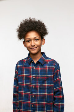 Vertical image of cute adorable school boy of 12 dressed in plaid shirt having funny fluffy afro haircut, standing against white studio wall, smiling, feeling good and happy. Carefree childhood