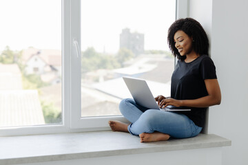 Young african woman sitting on the windowsill with a laptop.