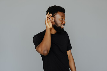 Close up portrait of surprised african man holding hand near ear and trying to hear the information isolated on gray background