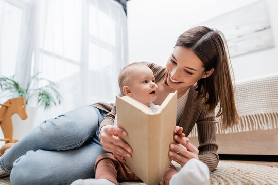 Young mother holding book near son on rug near bed at home.