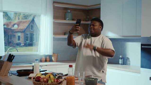 View of young obese African American man dancing on modern kitchen. Footage of guy in white t-shirt holding smartphone in hand, making video for social networks. Early morning. Indoors.