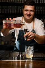smiling male bartender pouring alcoholic drink from crystal bottle into steel jigger