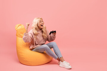 Overjoyed female gamer with joystick making YES gesture, feeling happy over her victory, sitting in beanbag chair © Prostock-studio