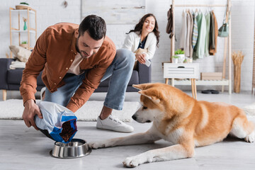 happy man adding pet food in bowl near akita inu dog and blurred girlfriend.