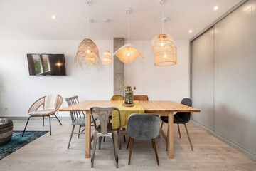 Living room with a wooden dining table, upholstered wooden and metal chairs and wicker lamps, a light wooden floor and a built-in wardrobe with gray sliding doors