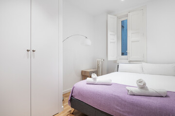 Bedroom with double bed with purple blanket and rolled towels, built-in wardrobe and windows with wooden shutters