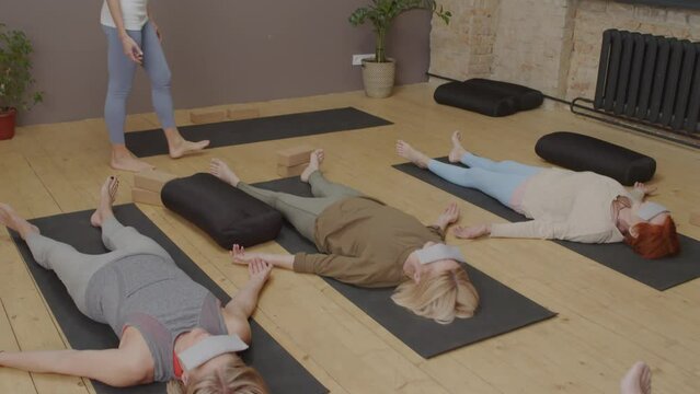 Tracking of aged women and men lying on backs on floor in modern yoga studio, meditating, young female fitness instructor walking around and talking
