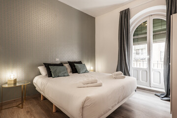 bedroom with king size bed, green and black cushions, clothespin and wall with decorative paper, wooden floor and circular bedside tables, balcony with wooden doors with semicircular arch