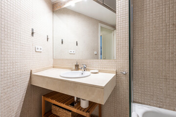 Bathroom with white porcelain sink on cream marble and mosaic countertop, frameless mirror set into the wall and wooden cabinet below the counter