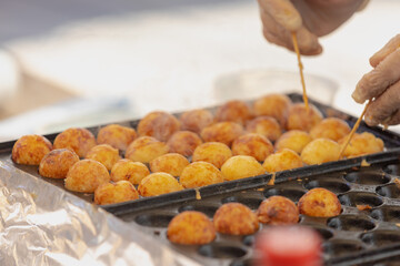 Selective focus of golden yellow Takoyaki balls cooking in a special molded pan at market stall with blurred hand while making, Japanese snack made of a wheat flour-based batter, Delicious street food