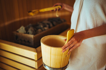 Beautiful woman adding steam at the sauna. Young beautiful woman enjoying the sauna at a beauty spa. Finnish wooden sauna bucket.