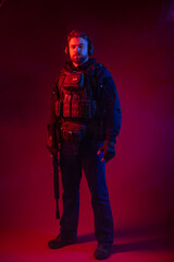 Fototapeta premium a fighter in equipment, body armor, headphones holds a machine gun, aims at the target, hits the target through the sight. military business concept. red background. blue-red light