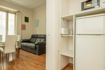 Fototapeta na wymiar Pantry with refrigerator next to a living room with a two-seater sofa and a glass dining table with white chairs