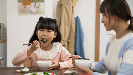closeup of cute asian girl enjoying eating healthy green vegetable with chopsticks at dining table. she smiles at her mother as she is chewing the food